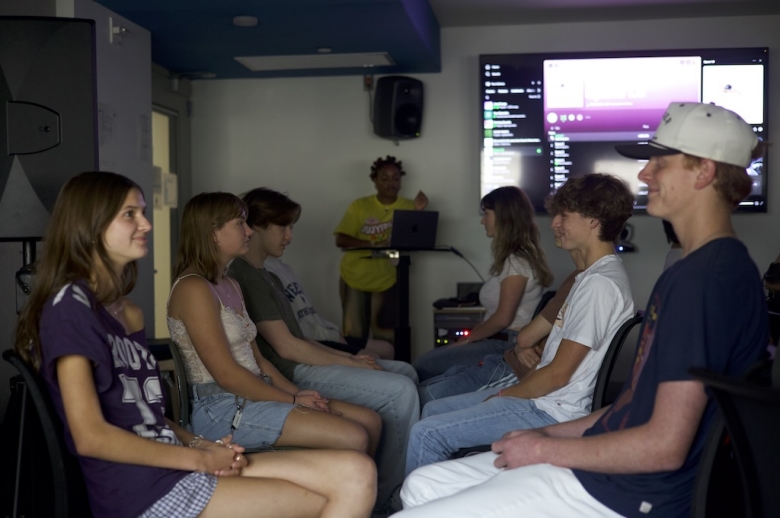 Tisch Summer High School students sit in pairs facing each other during a class at the Clive Davis Institute of Recorded Music. Behind them, the class instructor stands at a podium with a laptop presenting at the front of the classroom.